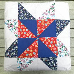 May include: A white cushion with a blue and red star design. The star is made of fabric squares with different floral patterns. The cushion is ideal for adding a touch of country charm to any room.