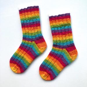 May include: A pair of rainbow-striped knitted socks. The socks display bands of red, orange, yellow, green, blue, and purple. The cuffs have a ruffled edge. The socks are made of a soft, textured yarn. The socks are suitable for a range of foot sizes.
