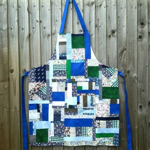 May include: A blue patchwork apron made from a variety of fabrics in shades of blue, green, and white. The apron has a large pocket in the front and adjustable straps.