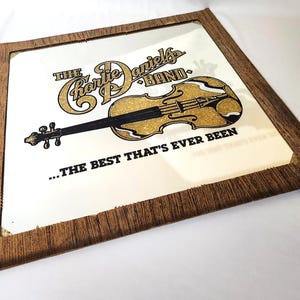 May include: A square mirror with a brown wood-grain frame featuring a gold glitter graphic of a violin and the text THE CHARLIE DANIELS BAND and ...THE BEST THAT'S EVER BEEN printed in black and gold across the reflective surface.