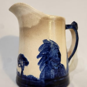 May include: A ceramic pitcher with a cream-colored base and a dark blue hand-painted design featuring a silhouette of a person with a feathered headdress and a tree. The pitcher has a wide pouring spout, a curved handle, and a dark blue base trim.
