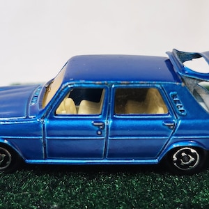 May include: A die-cast blue toy car with an open trunk. The vintage vehicle has a metallic finish, black wheels, and cream-colored interior. The car is set against a green, textured surface.