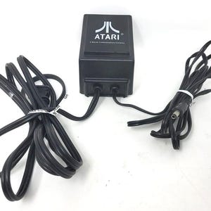 May include: Black Atari power supply with the Atari logo and text. The power supply has two black cords attached, one coiled and the other bundled with a white tie. The power supply is on a white surface.