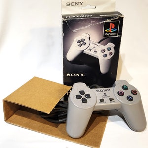 May include: A gray Sony PlayStation controller with a black cord and a gray box with the Sony PlayStation logo. The controller has a black directional pad, four black buttons, and two black triggers.