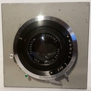 May include: A vintage camera lens assembly with a silver-colored metal body. The lens has a black circular aperture with markings including "FOCUS 7 IN." and "GOERZ DAGOR." The lens also has a "SYNCHRO-COMPUR" dial and various f-stop settings.