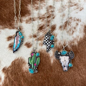 May include: Five silver necklaces with different pendants. The pendants are a turquoise feather, a checkered lightning bolt, a green cactus, and a white skull with turquoise accents.