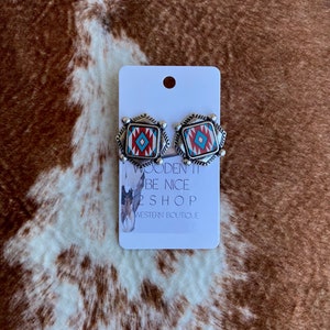May include: Silver-toned square stud earrings with a Southwestern design. The earrings feature a red, white, and turquoise geometric pattern. The earrings are displayed on a white card with the text "WOODEN IT BE NICE 2 SHOP WESTERN BOUTIQUE".