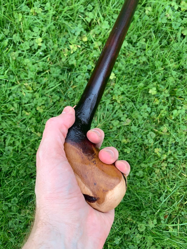 May include: A wooden walking stick with a dark brown stain and a natural wood handle. The handle is shaped to fit comfortably in the hand.