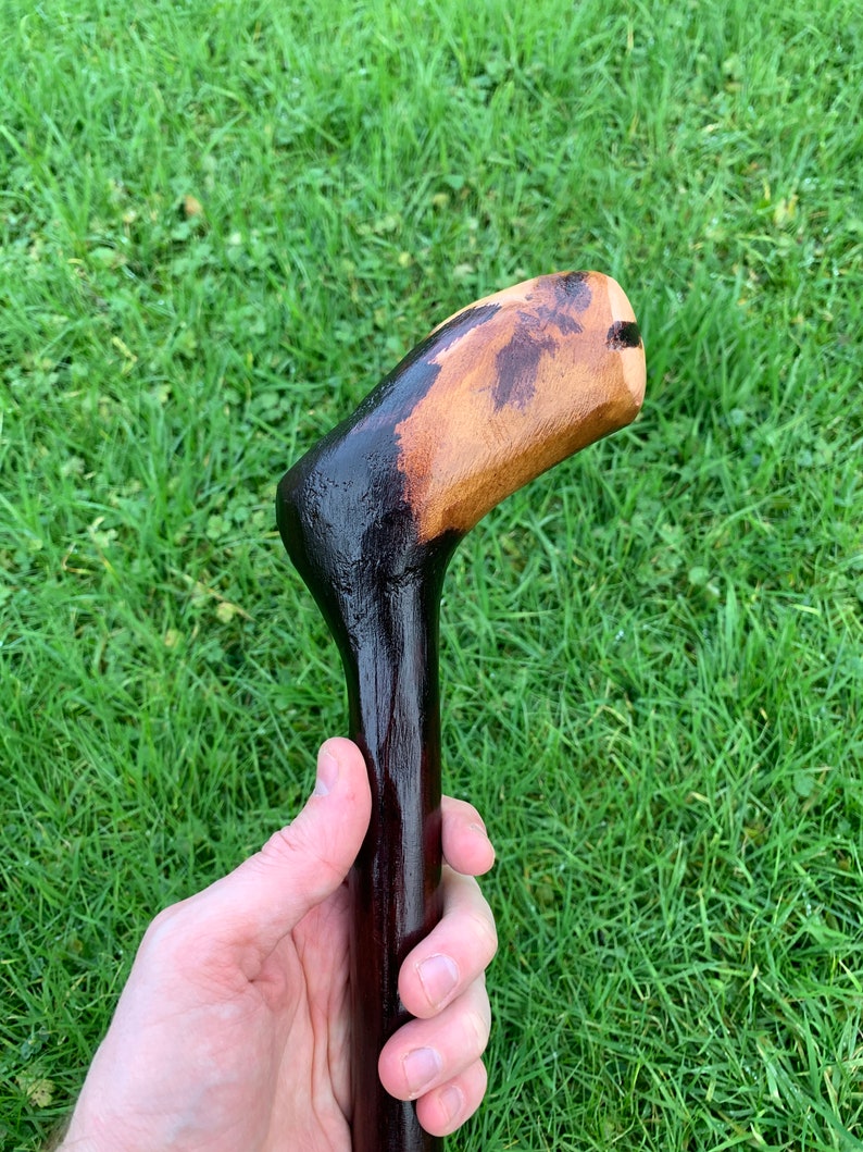 May include: A wooden walking stick with a dark brown stain and a natural wood handle. The handle is shaped like a curved hook.