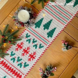 May include: A white crocheted table runner with a red and green Christmas tree design. The runner has a scalloped edge and is decorated with snowflakes and trees.