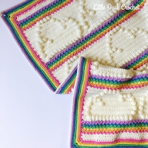 May include: Cream-coloured crocheted blanket with a rainbow border and heart-shaped patterns. The blanket has a vibrant border with pink, yellow, green, blue, and purple stripes. The text "Little Duck Crochet" is visible in the upper right corner.