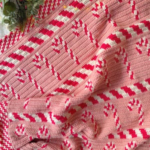 May include: A handmade pink crochet blanket with a candy cane pattern in red and white. The blanket features a repeating design of candy canes and striped accents, perfect for holiday decor. The edges are finished with a decorative border.