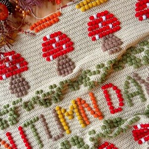 Puede incluir: Una manta de crochet con fondo beige y un dise&ntilde;o colorido. La manta presenta la palabra "AUTUMN" en hilo naranja, amarillo y verde. Tambi&eacute;n hay setas rojas y blancas con tallos marrones.