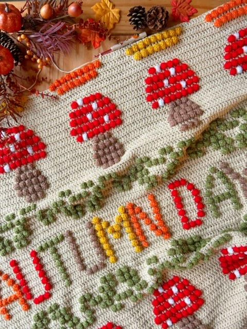 Puede incluir: Una manta de crochet con fondo beige y un dise&ntilde;o colorido. La manta presenta la palabra "AUTUMN" en hilo naranja, amarillo y verde. Tambi&eacute;n hay setas rojas y blancas con tallos marrones.