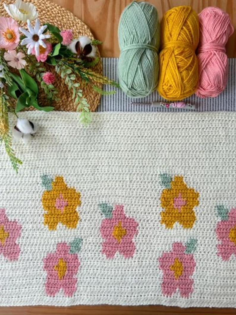 May include: A white crocheted blanket with a floral design. The blanket features yellow and pink flowers with green leaves. Three balls of yarn are visible in the background, one green, one yellow, and one pink.