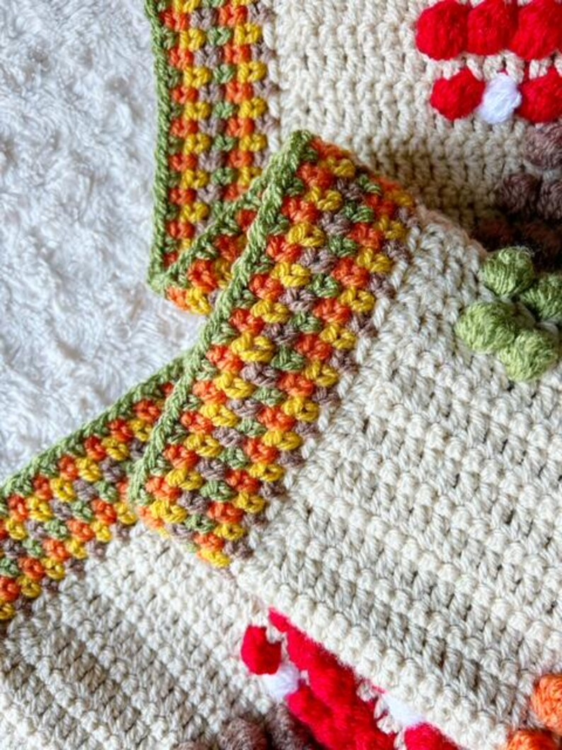 Puede incluir: Primer plano de una manta de ganchillo con un borde multicolor. El borde est&aacute; hecho de hilo verde, naranja, amarillo y marr&oacute;n. La manta est&aacute; hecha de hilo de color crema.