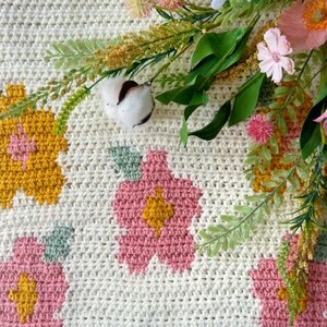 May include: A white crocheted blanket with pink and yellow flowers. The blanket is decorated with crocheted flowers in shades of pink and yellow.