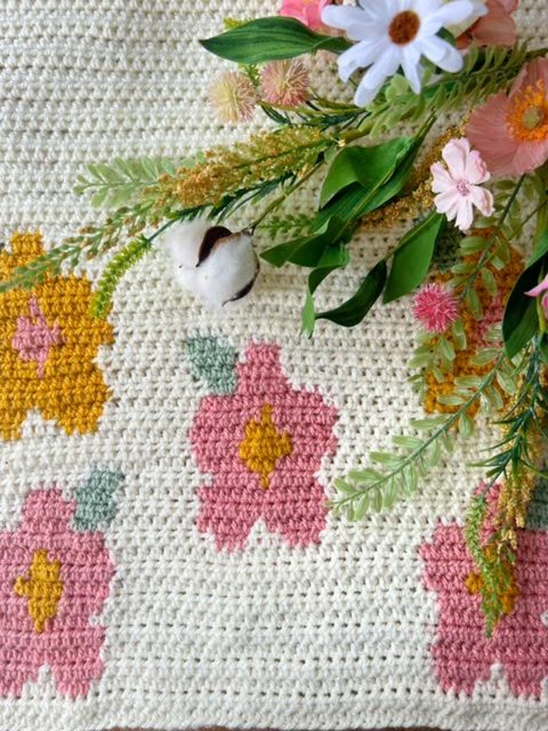 May include: A white crocheted blanket with pink and yellow flowers. The blanket is decorated with crocheted flowers in shades of pink and yellow.