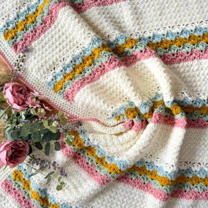 May include: A crocheted blanket featuring alternating horizontal stripes of cream, mustard yellow, light blue, and dusty pink. The texture consists of solid cream sections interspersed with decorative shell-stitch bands in the colored yarn.