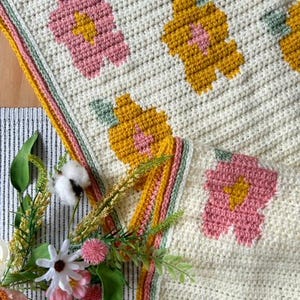 May include: A crocheted blanket with a floral pattern in shades of pink, yellow, and green. The blanket has a white background and a border in shades of yellow, pink, and green.