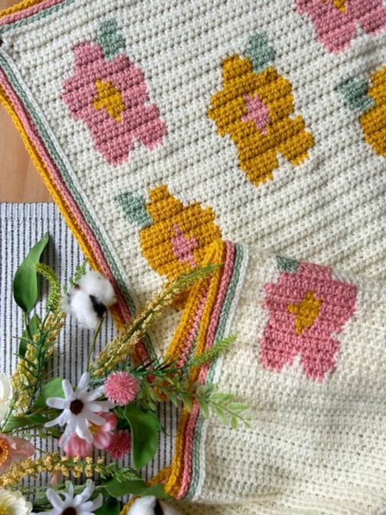 May include: A crocheted blanket with a floral pattern in shades of pink, yellow, and green. The blanket has a white background and a border in shades of yellow, pink, and green.