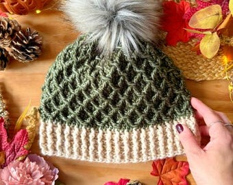 May include: A green and beige knitted beanie with a faux fur pom-pom. The hat features a textured diamond pattern on the crown and a ribbed brim. The pom-pom is gray and fluffy. The hat is surrounded by fall-themed decorations, including leaves and flowers.