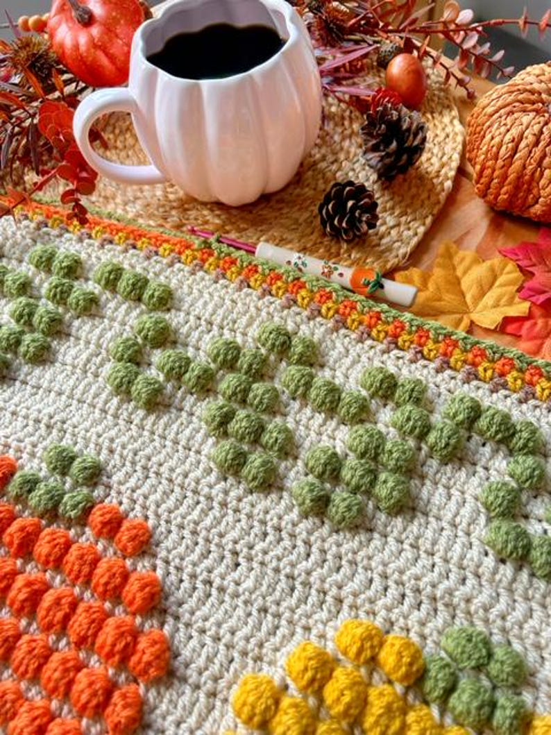 Puede incluir: Una manta de ganchillo con un patr&oacute;n texturizado de c&iacute;rculos naranja, amarillo y verde. La manta est&aacute; hecha con hilo beige y tiene un borde de hilo marr&oacute;n, naranja y amarillo.