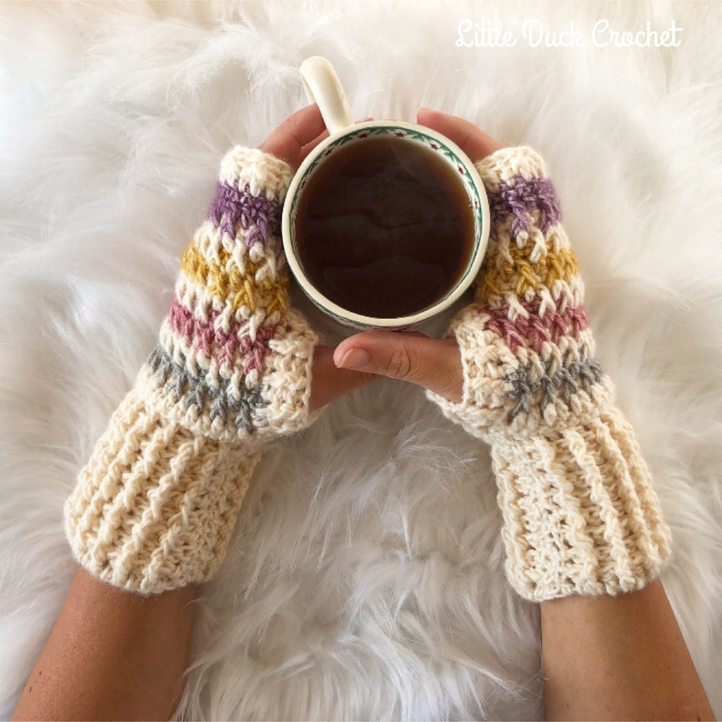 May include: A pair of crocheted fingerless gloves with a colorful striped pattern in shades of pink, purple, yellow, and gray. The gloves are made with a cream-colored yarn and are being worn by a person holding a cup of tea.