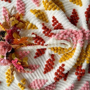 May include: A cream-colored, crocheted blanket with a textured pattern of raised, leaf-like shapes in shades of red, pink, and yellow. The blanket is folded, revealing the intricate stitchwork and color variations. Decorative flowers and foliage are arranged on the blanket.