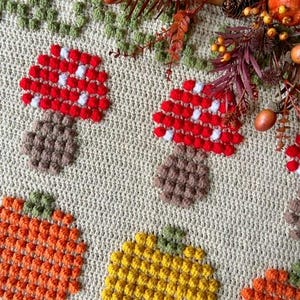 Puede incluir: Una manta de ganchillo con tem&aacute;tica oto&ntilde;al con setas, calabazas y hojas. La manta est&aacute; hecha con una variedad de colores, incluyendo rojo, naranja, amarillo, verde y marr&oacute;n.