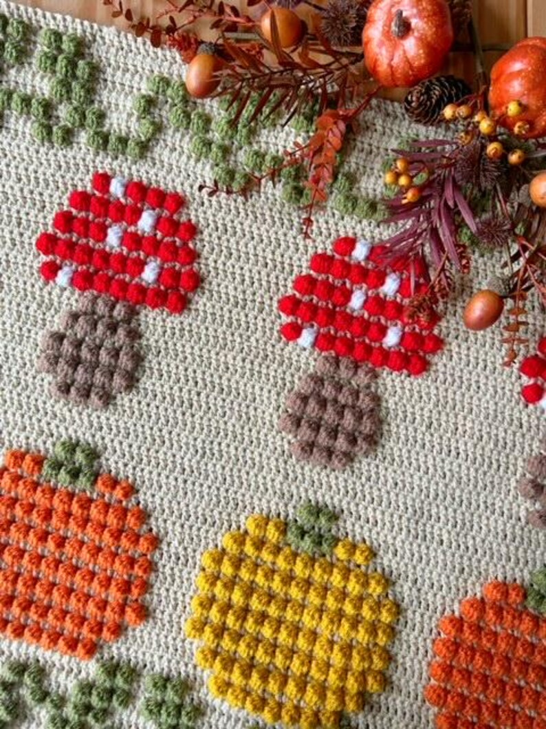 Puede incluir: Una manta de ganchillo con tem&aacute;tica oto&ntilde;al con setas, calabazas y hojas. La manta est&aacute; hecha con una variedad de colores, incluyendo rojo, naranja, amarillo, verde y marr&oacute;n.