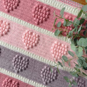 Pode incluir: Cobertor de crochê feito à mão com um padrão de coração em relevo. Apresenta listras rosa, rosa claro, branco e roxo. Os corações texturizados adicionam dimensão. Feito de fio macio, ideal para um ambiente acolhedor.