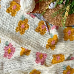 May include: A crocheted blanket with a floral pattern in shades of yellow, pink, and green. The blanket is made with a white yarn and has a textured surface.
