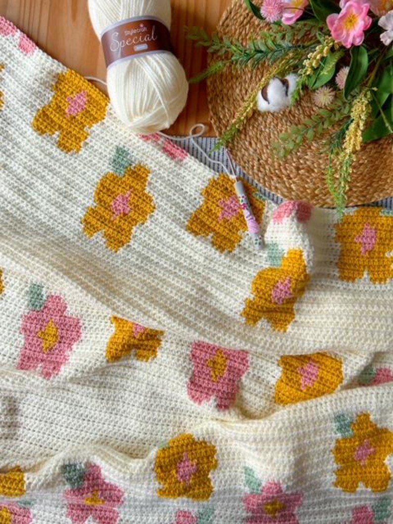May include: A crocheted blanket with a floral pattern in shades of yellow, pink, and green. The blanket is made with a white yarn and has a textured surface.