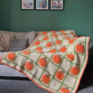 May include: A crocheted blanket featuring a grid pattern of cream-colored squares separated by light green textured lines. Each square contains a raised, orange, bobble-stitch pumpkin motif with a small brown stem at the top, finished with an orange border.