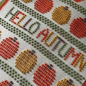 May include: A crocheted blanket with a white background and a pattern of orange and yellow pumpkins. The blanket has the words "Hello Autumn" written in orange yarn.