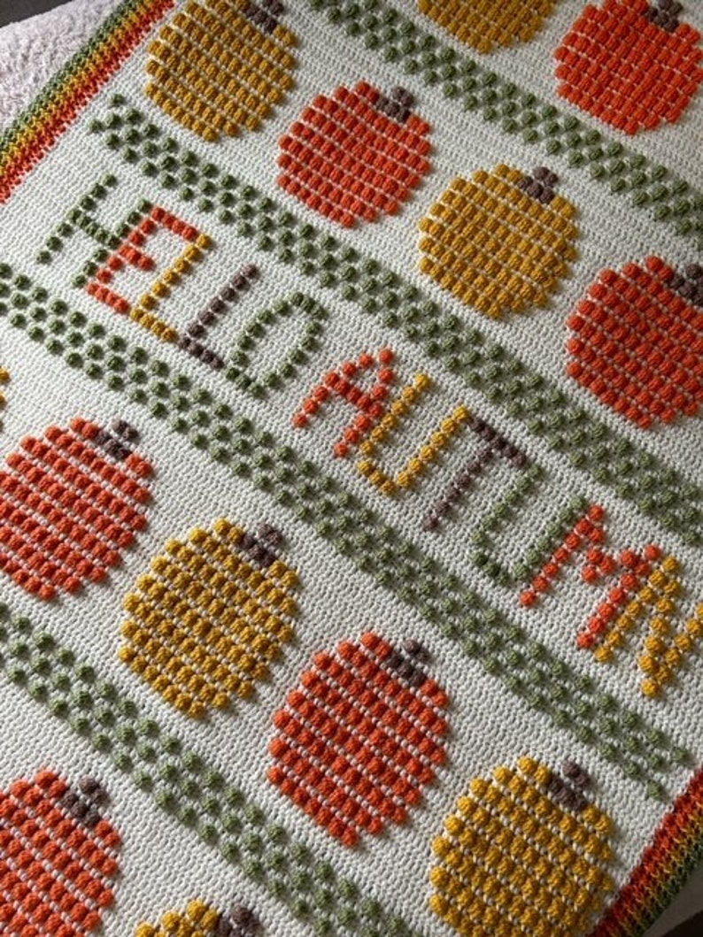 May include: A crocheted blanket with a white background and a pattern of orange and yellow pumpkins. The blanket has the words "Hello Autumn" written in orange yarn.