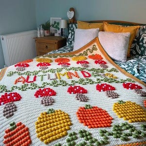 Puede incluir: Una manta de crochet con fondo crema y un tema oto&ntilde;al colorido. La manta presenta setas, calabazas y hojas en tonos rojos, naranjas, amarillos y verdes. El texto "Oto&ntilde;o" est&aacute; cosido en hilo rojo a trav&eacute;s del centro de la manta.