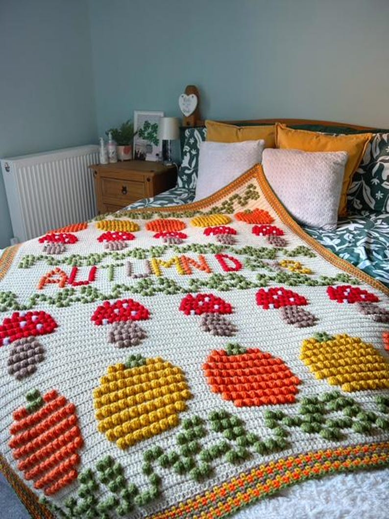 Puede incluir: Una manta de crochet con fondo crema y un tema oto&ntilde;al colorido. La manta presenta setas, calabazas y hojas en tonos rojos, naranjas, amarillos y verdes. El texto "Oto&ntilde;o" est&aacute; cosido en hilo rojo a trav&eacute;s del centro de la manta.