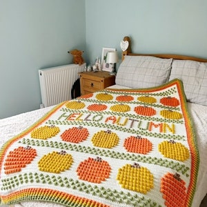 May include: A white crocheted blanket with a green, orange, and yellow pattern. The blanket features the words "Hello Autumn" in orange and yellow yarn.