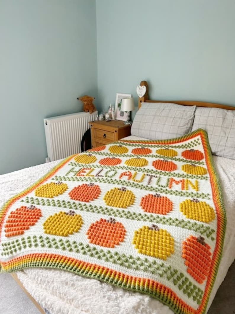 May include: A white crocheted blanket with a green, orange, and yellow pattern. The blanket features the words "Hello Autumn" in orange and yellow yarn.