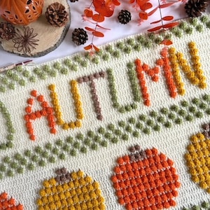 May include: A crocheted blanket with the word "AUTUMN" in orange, yellow, and brown yarn. The blanket features a repeating pattern of pumpkins and leaves in various shades of green, orange, and yellow.