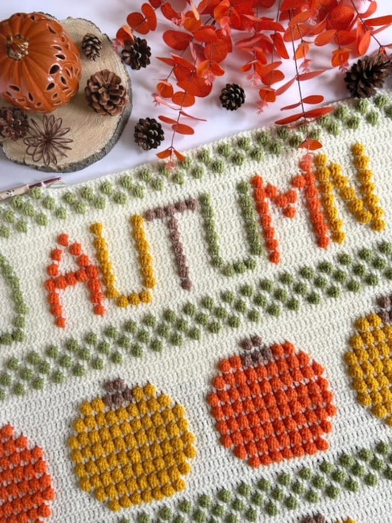 May include: A crocheted blanket with the word "AUTUMN" in orange, yellow, and brown yarn. The blanket features a repeating pattern of pumpkins and leaves in various shades of green, orange, and yellow.