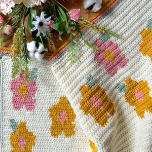 May include: A crocheted blanket with a white background and a floral pattern in shades of pink, yellow, and green. The blanket is folded in half and has a border of white and green yarn.