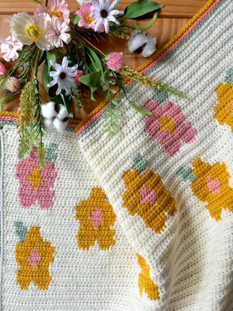 May include: A crocheted blanket with a white background and a floral pattern in shades of pink, yellow, and green. The blanket is folded in half and has a border of white and green yarn.