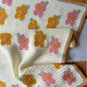 May include: A crocheted blanket with a white background and a pattern of pink and yellow flowers. The blanket has a light green, white, and yellow border.