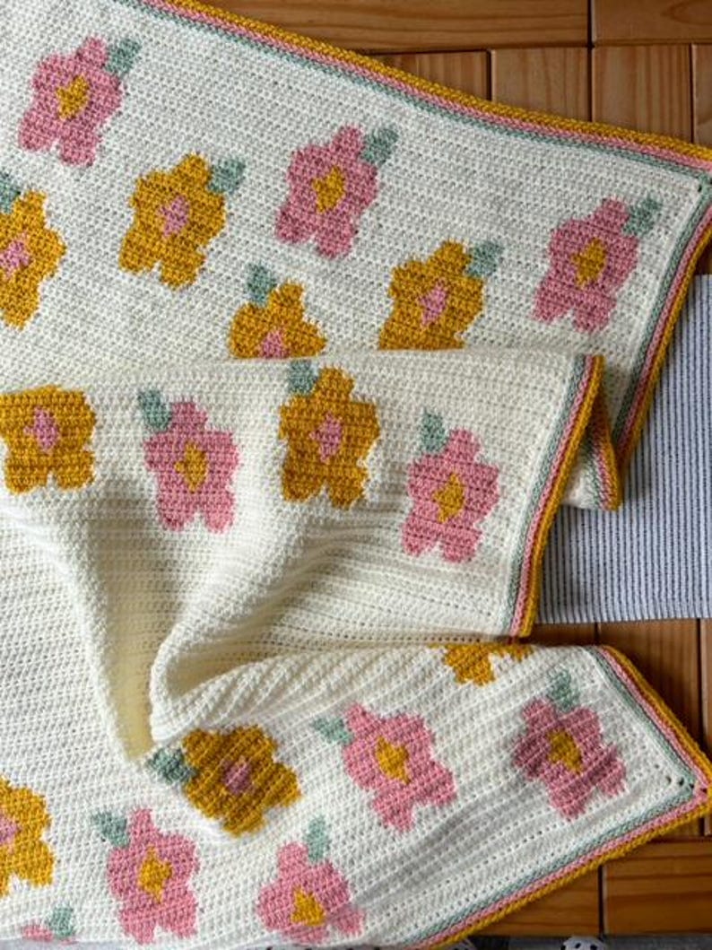 May include: A crocheted blanket with a white background and a pattern of pink and yellow flowers. The blanket has a light green, white, and yellow border.
