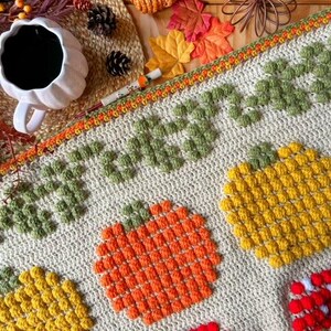 Puede incluir: Una manta de ganchillo con tem&aacute;tica oto&ntilde;al con calabazas, hojas y bellotas. La manta est&aacute; hecha con una variedad de colores, incluyendo naranja, amarillo, verde y marr&oacute;n. La manta es perfecta para agregar un toque de oto&ntilde;o a la decoraci&oacute;n de tu hogar.