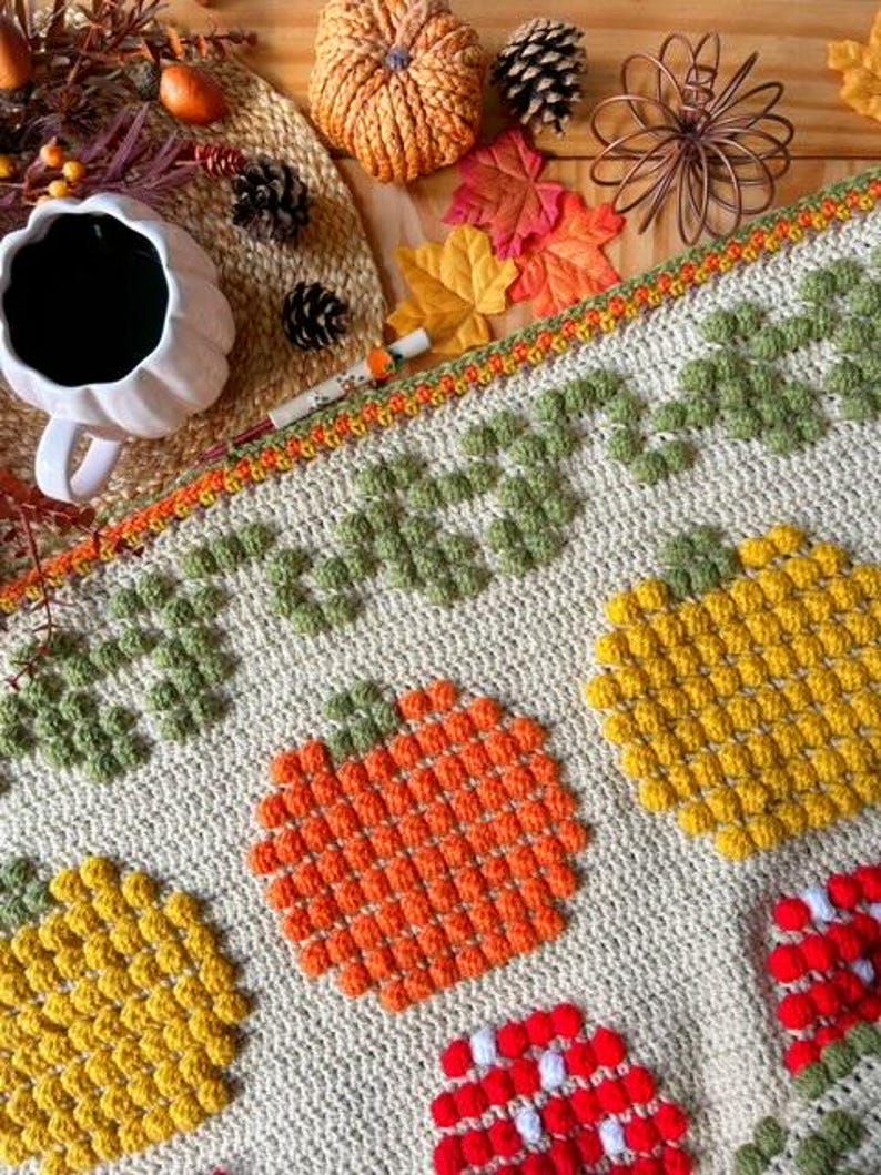 Puede incluir: Una manta de ganchillo con tem&aacute;tica oto&ntilde;al con calabazas, hojas y bellotas. La manta est&aacute; hecha con una variedad de colores, incluyendo naranja, amarillo, verde y marr&oacute;n. La manta es perfecta para agregar un toque de oto&ntilde;o a la decoraci&oacute;n de tu hogar.