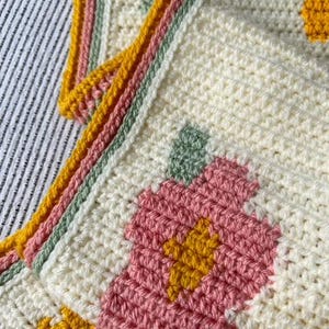 May include: A crocheted blanket with a floral pattern in shades of pink, yellow, and green. The blanket has a white background and a border of yellow, pink, and green.